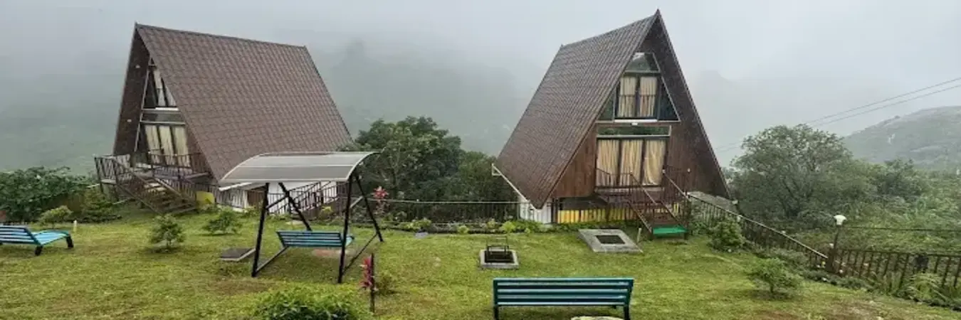 A-frame wooden resort cottages with lawn seating set against misty hills at JMount Vagamon Resort in Kannamkulam