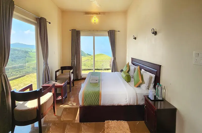 Luxury resort bedroom featuring a plush double bed and large glass windows overlooking green Vagamon hills