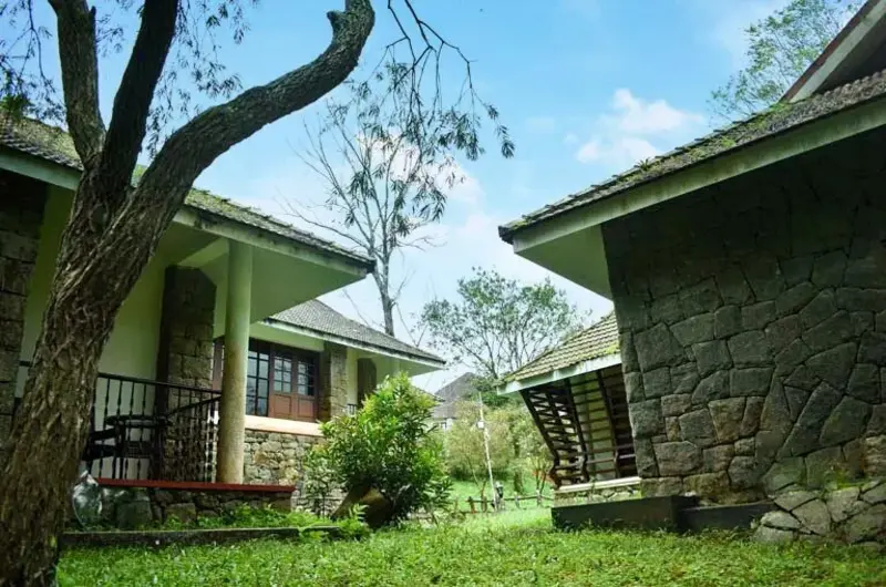 Exterior view of an eco-cottage villa surrounded by lush green lawns and tropical trees at Adrak Summer Sand Hill Resort Vagamon