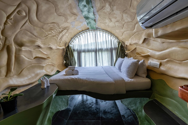 Unique cave-style guest room with wall reliefs and curved window facing green landscapes in Vagamon
