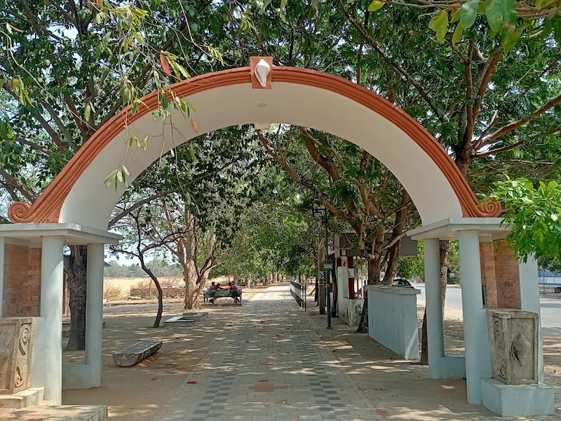 Tree-lined paved walkway viewed through an arched stone gateway on ashram grounds in Kerala
